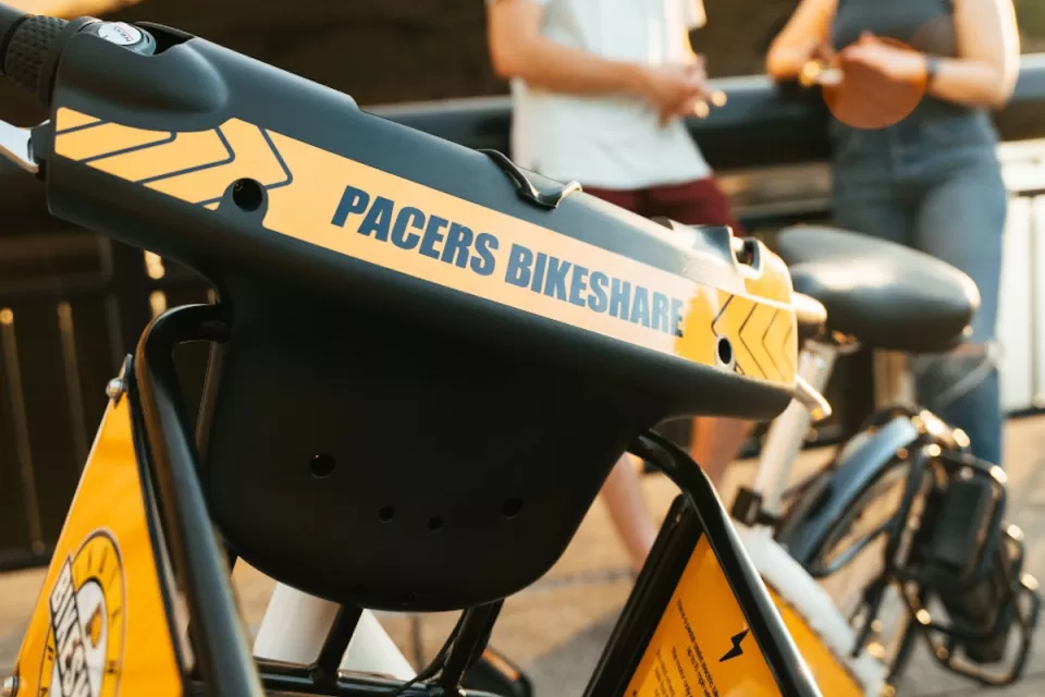 Close-up of yellow Pacers Bikeshare e-bike parked outdoors with blurred people in background.